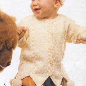 May include: A smiling baby wearing a cream-colored knitted outfit with a matching hat. The outfit includes a button-down cardigan with a unique jagged hemline adorned with white pom-poms. The baby is holding a stuffed animal.
