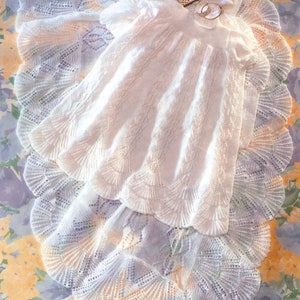 May include: A white crocheted baby blanket with a delicate lace pattern. The blanket is folded in half and has a small card with a baby's picture on it.