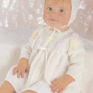 May include: A white knitted baby dress with a white satin ribbon and flower embellishments. The dress has long sleeves and a round neckline. The baby is wearing a white knitted hat with flower embellishments.