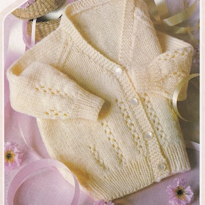 Könnte beinhalten: Ein cremefarbener Strickcardigan mit V-Ausschnitt und Knopfleiste. Der Cardigan hat ein zartes Spitzenmuster an den Ärmeln und vorne.