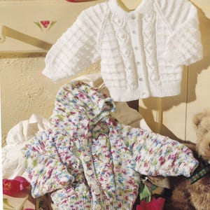 Op de afbeelding: Twee babyvesten. Het bovenste vest is wit met een textuurpatroon en knoopsluiting. Het onderste vest is veelkleurig met een capuchon en knoopsluiting. Gebreid.