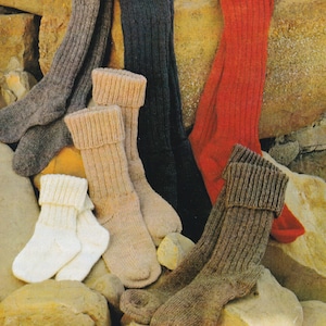 May include: A collection of six knitted socks in various colors and styles, including brown, gray, white, black, and red. The socks are arranged on a background of rocks.