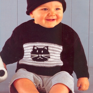 Könnte beinhalten: Ein Baby trägt einen schwarzen Pullover mit einem weißen und grauen gestreiften Katzendesign auf der Vorderseite. Das Baby trägt auch eine schwarze Strickmütze und graue Shorts.