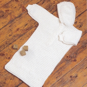 Puede incluir: Bata de bebé de ganchillo blanco con capucha a juego. La bata está colocada sobre una superficie de madera con tres bloques de madera.