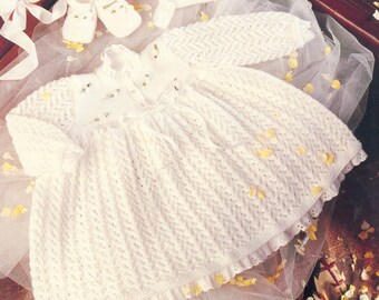 May include: A white knitted baby dress with a lace trim and a bow at the waist. The dress is laid out on a white tulle fabric with yellow flower petals scattered around it.