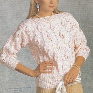 Può includere: Un maglione bianco lavorato a maglia con un motivo a treccia e macchie rosa. Il maglione ha un collo a barca e maniche lunghe.