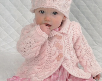 May include: A pink knitted baby cardigan with a cable knit pattern and buttons. The cardigan is worn over a pink and white plaid dress. The baby is wearing a matching pink knitted hat.
