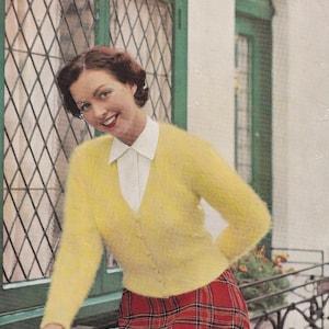 May include: A woman wearing a yellow fuzzy cardigan sweater with a white collared shirt underneath. She is wearing a red and black plaid skirt and is standing next to a window.