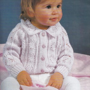 May include: A white knitted baby cardigan with a lace pattern and button closure. The cardigan has a collar and long sleeves.