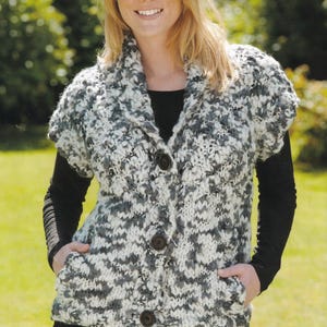 May include: A short-sleeved, button-up cardigan in a chunky knit pattern. The sweater features a mix of white and gray yarns, creating a textured appearance. It has a shawl collar and three dark brown buttons. The person is wearing a black shirt underneath.