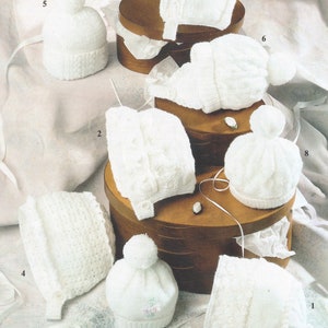 Könnte beinhalten: Acht weiße gestrickte Baby-Mützen mit verschiedenen Designs und Texturen. Die Mützen werden auf Holzkisten und einer weißen Oberfläche präsentiert. Die Zahlen 1 bis 8 sind auf dem Bild sichtbar.