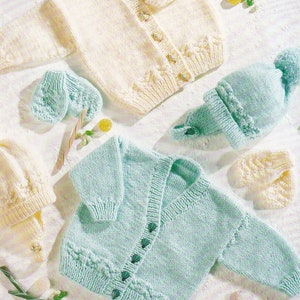 May include: Two knitted baby cardigans, one in cream and one in light blue, with matching hats and mittens. The cardigans have button fastenings and are made with a simple stitch pattern.
