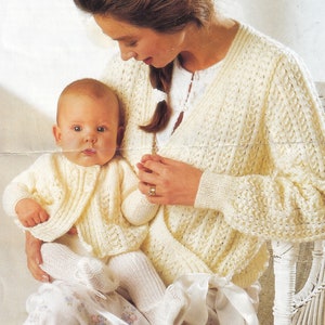 May include: A woman and a baby are wearing matching white knitted cardigans with a lacy pattern. The woman is holding the baby in her lap. The baby is wearing white socks.