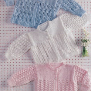 Op de afbeelding: Drie gebreide baby-vestjes in blauw, wit en roze met een knoopsluiting en een delicaat kantpatroon.