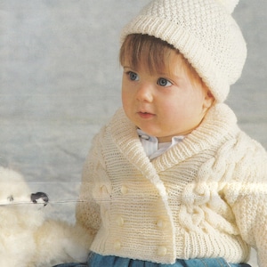 Könnte beinhalten: Ein weißer Strickcardigan mit Zopfmuster und Knopfleiste. Der Cardigan wird von einem Baby getragen, das eine weiße Strickmütze mit Bommel trägt.