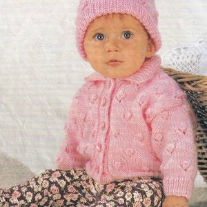 May include: A pink knitted baby cardigan with a button fastening and a matching pink knitted hat with a pom pom on top. The cardigan features a heart-shaped stitch pattern.