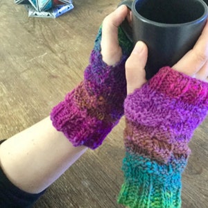 Könnte beinhalten: Ein Paar bunte gestrickte Fingerhandschuhe mit einem Farbmuster in Lila, Grün und Braun. Die Handschuhe werden von einer Person getragen, die eine schwarze Tasse hält.