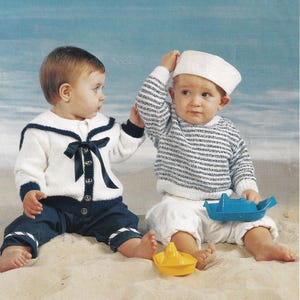 May include: Two toddlers wearing sailor-themed knitted sweaters play with toy boats on a sandy beach. One toddler is wearing a white sweater with a blue collar and a black bow. The other toddler is wearing a white and blue striped sweater and a white sailor hat.