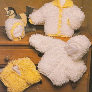 Könnte beinhalten: Drei gestrickte Baby-Pullover in Weiß und Gelb. Die Pullover sind aus einem flauschigen Garn gefertigt und haben einen Knopfverschluss. Die Pullover werden auf einer Holzoberfläche präsentiert.
