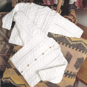 以下が含まれることがあります： 繊細なレース模様とボタン留めが施された、白のクロシェ編みベビーセーターとカーディガンセットです。