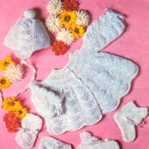 Puede incluir: Un conjunto de bebé blanco tejido, que incluye una chaqueta, gorro, manoplas y patucos. El conjunto presenta un patrón texturizado y está adornado con pequeños lazos. Los artículos están dispuestos sobre un fondo rosa con flores coloridas.