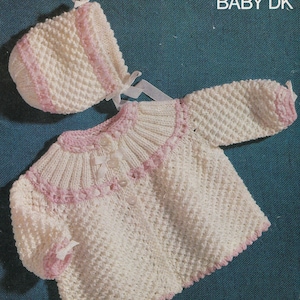 May include: A white knitted baby jacket with pink trim and buttons, and a matching white knitted hat with pink trim. The jacket and hat are made with a textured stitch pattern. The text "BABY DK" is printed in the top right corner of the image.