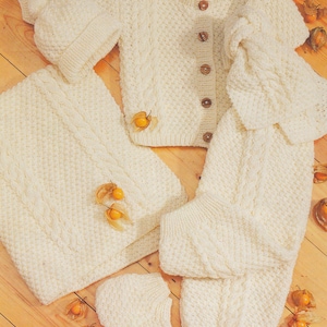 Könnte beinhalten: Ein weißes Strick-Baby-Outfit-Set bestehend aus einem Cardigan, einer Hose, einer Mütze und Booties. Das Set hat ein Zopfmuster und ist aus weichem Garn gefertigt.