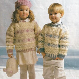 Puede incluir: Dos niños con suéteres de punto con un patrón geométrico. La niña lleva un gorro rosa y sostiene un animal de peluche blanco. El niño lleva un suéter azul y blanco.