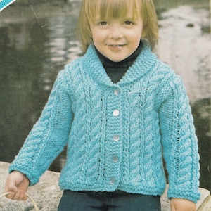 May include: A light blue cable-knit cardigan with a shawl collar and three silver buttons. The sleeves and front panels feature a textured cable pattern. The cardigan is worn over a black turtleneck.
