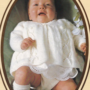 Może przedstawiać: Biały dzianinowy kardigan dla niemowlęcia z wzorem warkocza i zapięciem na guziki. Kardigan jest noszony przez niemowlę z białą koronkową lamówką na dole odzieży.