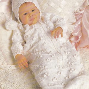 Puede incluir: Un saco de dormir de bebé blanco tejido con un gorro blanco tejido. El saco de dormir tiene una cremallera en la parte delantera y un patrón texturizado con pequeños pompones blancos.