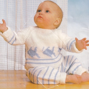 May include: A white knitted baby onesie with blue stripes and fish designs. The onesie is being worn by a baby sitting on a wooden floor.