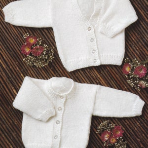 May include: Two white knitted baby cardigans with button fastenings. The cardigans are laid out on a brown woven background.