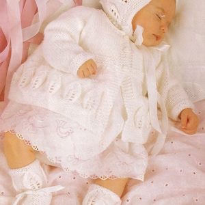 Puede incluir: Un bebé dormido con un conjunto de punto blanco, que incluye un gorro, una rebeca, un vestido y patucos. El conjunto presenta recortes en forma de hoja y está atado con cintas blancas. El fondo es de tela rosa suave.