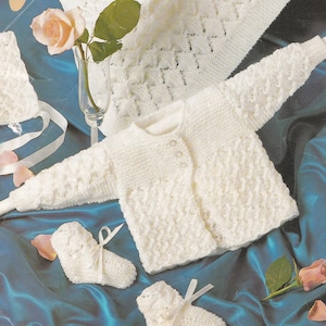 May include: A white knitted baby cardigan, booties, and a blanket. The cardigan has a button fastening and a delicate stitch pattern. The booties have white satin ribbons. The blanket has a simple, open stitch pattern.