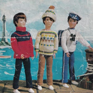 May include: Three knitted dolls wearing different outfits. The doll on the left is wearing a red and blue striped jumper, blue trousers, and white shoes. The doll in the middle is wearing a cream, yellow, and brown patterned jumper, brown trousers, and white shoes. The doll on the right is wearing a white jumper with a blue sailor hat, blue trousers, and white shoes.