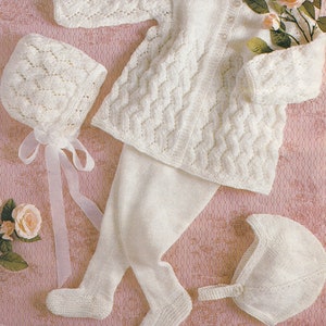 Puede incluir: Un conjunto de ropa de punto blanco para bebé que incluye un cárdigan, pantalones y dos gorros. El cárdigan tiene cierre de botón y un delicado patrón de encaje. Los pantalones son de punto liso y los gorros tienen un patrón de encaje y un detalle de botón.