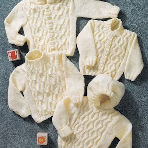 May include: Three white knitted baby sweaters with a cable knit pattern. The sweaters are laid out on a blue surface with wooden blocks featuring animal designs. The text on the blocks reads "36-61 cm 14-24 in".