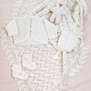 May include: A white knitted baby set including a cardigan, a hat, booties, and a blanket. The blanket has a lacy pattern and the set is laid out on a pink surface.