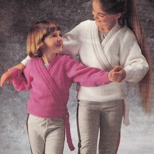 May include: Two girls wearing matching wrap sweaters. The girl on the left is wearing a pink sweater and the girl on the right is wearing a white sweater. Both girls are wearing grey trousers with black stripes.