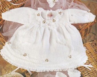 May include: A white knitted baby dress with a lace trim and floral embroidery. The dress is paired with a matching bonnet and booties. The set is displayed on a wicker basket with a pink tulle fabric.