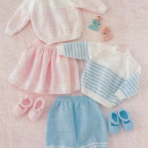 May include: A set of knitted baby clothes, including a pink skirt, a white and pink sweater, blue pants, and a blue and white striped sweater. The clothes are laid out on a pink background with white polka dots. The text "DOUBLE KNITTING" and "Sizes 16-22 in 41-56 cm" is visible in the top right corner of the image.
