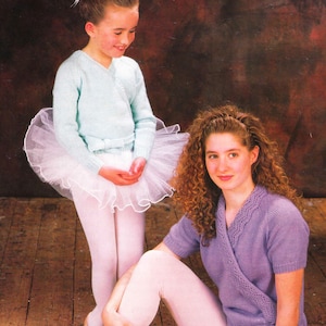 May include: Two young people in ballet outfits. One wears a pale blue wrap-around cardigan, a white tutu, and pink tights. The other wears a lilac wrap-around cardigan and pink tights. They are on a wooden floor with a brown background.