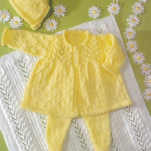 Puede incluir: Un conjunto de bebé de punto amarillo con un gorro a juego. El conjunto incluye un cárdigan, pantalones y un gorro. El conjunto está colocado sobre una manta de ganchillo blanca con un borde de adornos de margaritas blancas.