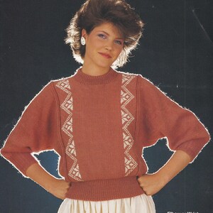 May include: A coral-coloured jumper with white geometric patterns on the front and sleeves. The jumper has a round neckline and elbow-length sleeves. The bottom of the jumper is ribbed. The text "Elbow or Wrist length sleeve" is visible in the image.