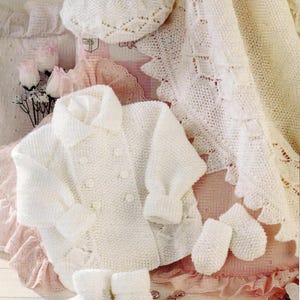 May include: A white knitted baby outfit set, including a coat, hat, booties, and mittens. The coat has a double-breasted design with buttons. The set is displayed on a pink surface with a floral background, showcasing a classic, handmade aesthetic.