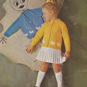 May include: A young girl wearing a yellow cardigan with white dog designs, a white pleated skirt, and white socks. She is holding a blue cardigan with white dog designs.