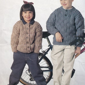 May include: Two children wearing knitted sweaters. A girl is wearing a brown cardigan with buttons and blue jeans. A boy is wearing a blue sweater with buttons and beige pants. Both children are standing next to a bicycle.