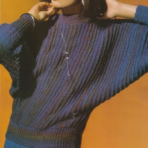 Könnte beinhalten: Ein lila-grüner Strickpullover mit hohem Kragen und langen Ärmeln. Der Pullover hat ein strukturiertes Strickmuster und wird von einer Person mit dunklen Haaren getragen.