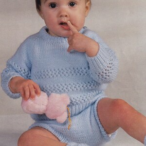 Op de afbeelding: Een baby in een lichtblauwe gebreide outfit, bestaande uit een trui, korte broek en sokken. De trui heeft een decoratief ajourpatroon over de borst en mouwen. De baby houdt een roze knuffel vast.